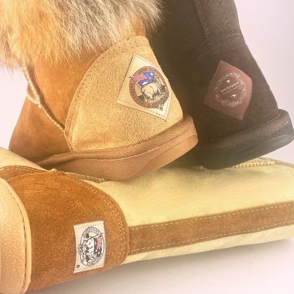 A close-up of several UGG-style boots showing the interior suede collar, fur trim, and interior lining. One boot has tan suede with a cream fur top and a patch logo, while another shows a dark brown suede boot and a third shows a cream-lined boot interior with a tan suede collar and a visible Sheepskins Downunder logo.